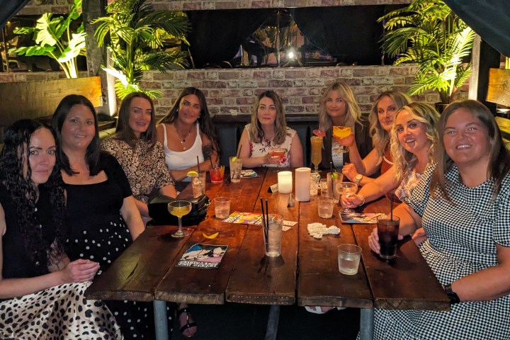 Group of ten women sitting at a dining table with drinks, surrounded by plants.