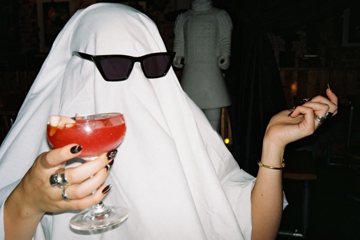 Person in ghost costume with sunglasses holding a cocktail.