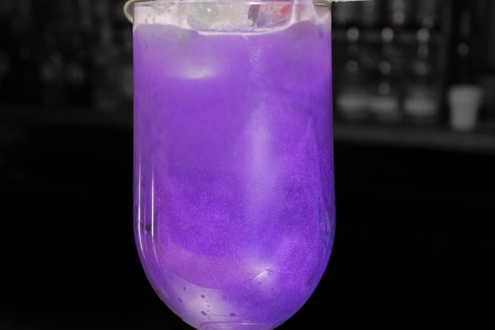 Purple Halloween cocktail in glass with fruit garnish on dark background.